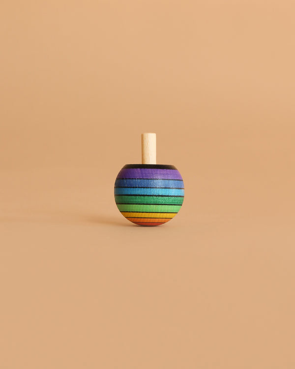 The Mader Rainbow Turn-Over Spinning Top - Red stands upright on a simple beige background, showcasing its vivid rainbow colors and wooden craftsmanship.