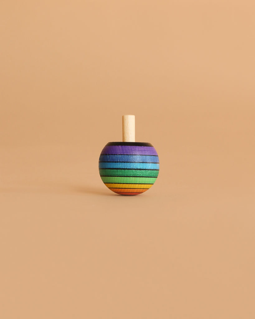 The Mader Rainbow Turn-Over Spinning Top - Red stands upright on a simple beige background, showcasing its vivid rainbow colors and wooden craftsmanship.