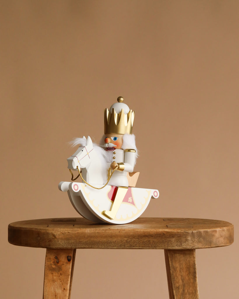 The Christian Ulbricht Collectible Wooden Nutcracker - Rider King wears a gold crown and white outfit, riding a white rocking horse, displayed on a rustic wooden stool against a plain beige background.