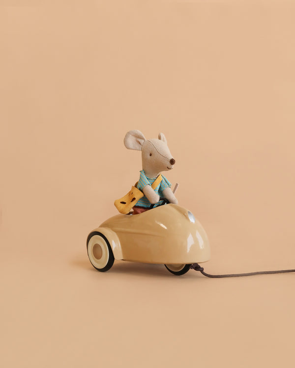 A Maileg Little Brother Adventure Set mouse, dressed in blue clothes, sits in a beige car holding a piece of yellow cheese against a plain peach background.