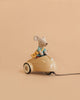 A Maileg Little Brother Adventure Set mouse, dressed in blue clothes, sits in a beige car holding a piece of yellow cheese against a plain peach background.