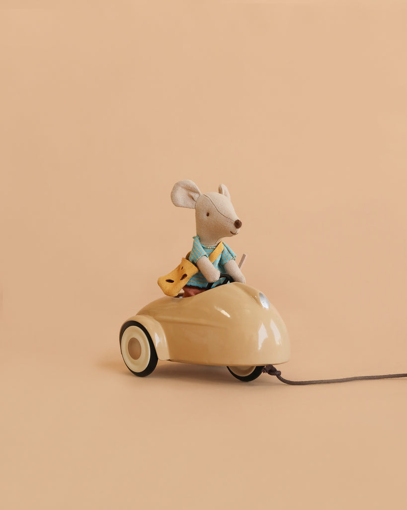 A Maileg Little Brother Adventure Set mouse, dressed in blue clothes, sits in a beige car holding a piece of yellow cheese against a plain peach background.