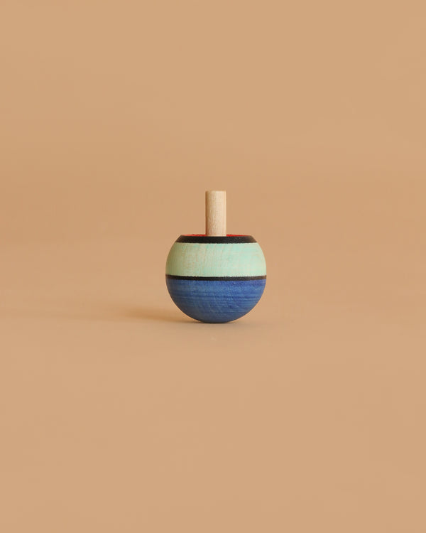 The Mader Confetti Turn-Over Spinning Top, made of maple wood with blue, mint green, and brown stripes, stands upright on a plain beige background.