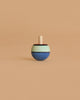 The Mader Confetti Turn-Over Spinning Top, made of maple wood with blue, mint green, and brown stripes, stands upright on a plain beige background.