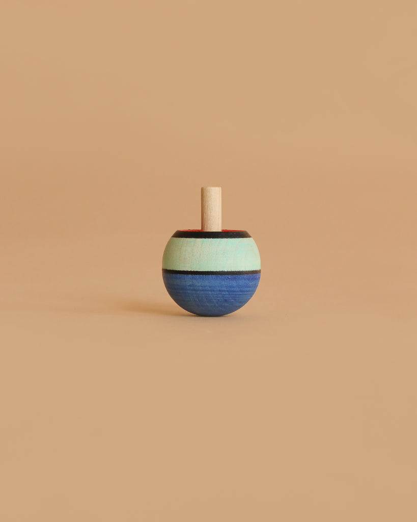 The Mader Confetti Turn-Over Spinning Top, made of maple wood with blue, mint green, and brown stripes, stands upright on a plain beige background.