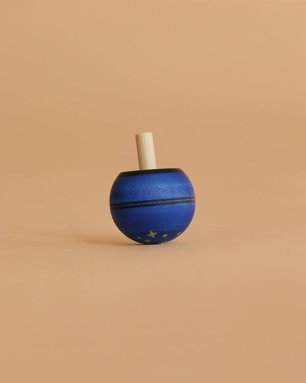 The Mader Luna Turn-Over Spinning Top, featuring a blue body, light-colored handle, and a delicate starry sky design, stands upright on a plain beige background, highlighting its whimsical charm and unique appeal.