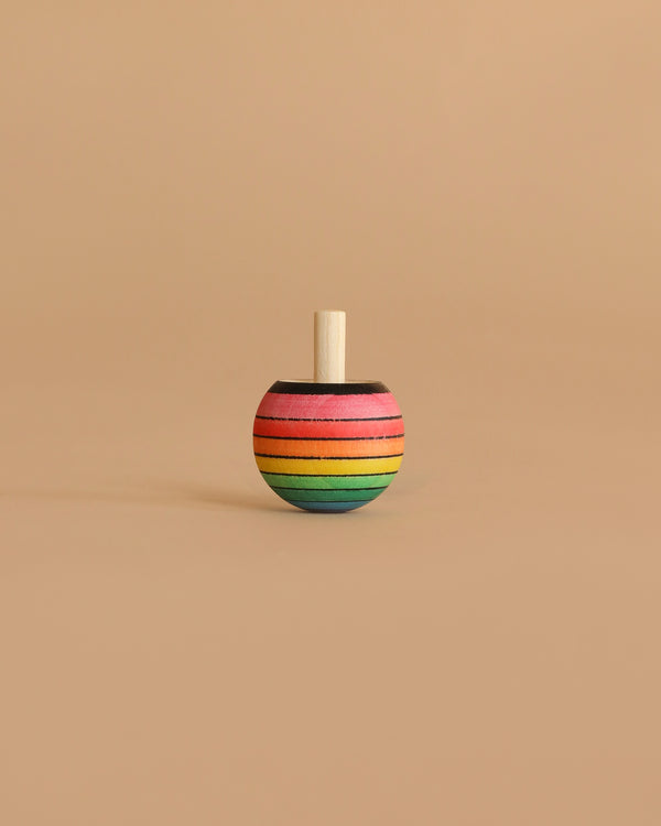 The Mader Rainbow Turn-Over Spinning Top - Purple, a wooden top featuring a vibrant rainbow gradient with purple accents, stands upright on a plain beige surface and background.