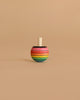 The Mader Rainbow Turn-Over Spinning Top - Purple, a wooden top featuring a vibrant rainbow gradient with purple accents, stands upright on a plain beige surface and background.