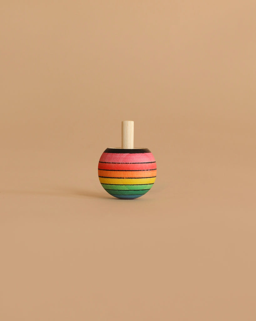 The Mader Rainbow Turn-Over Spinning Top - Purple, a wooden top featuring a vibrant rainbow gradient with purple accents, stands upright on a plain beige surface and background.