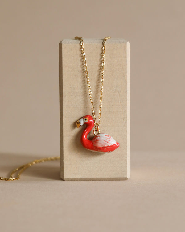 A red and white hand-painted porcelain Flamingo Necklace hangs from a gold chain draped over a beige block, set against a matching beige background.