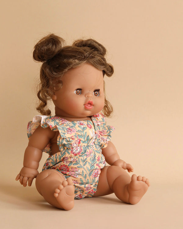 A Minikane Baby Doll With Sleeping Eyes (13") - Madeleine with light brown skin and curly brown hair styled in two buns sits against a beige background. The doll, anatomically correct and softly vanilla-scented, is dressed in a floral print romper with ruffled sleeves. Its facial expression is neutral, and its legs are extended forward.