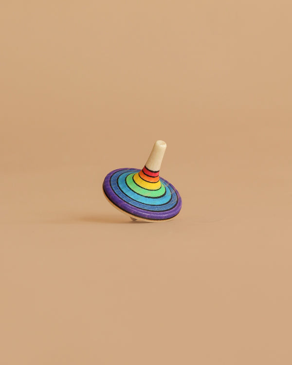 The Mader Rallye Striped Spinning Top, Small features concentric rainbow rings and a white stem, standing upright on a light brown surface with a matching background.