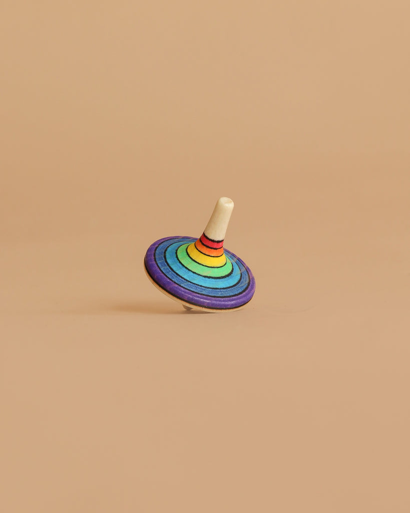 The Mader Rallye Striped Spinning Top, Small features concentric rainbow rings and a white stem, standing upright on a light brown surface with a matching background.