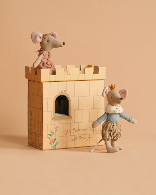 Two toy mice, dressed as prince and princess, are posed with the Maileg Princess And The Pea With Prince Set. One stands inside the floral cardboard castle box reading "The Princess Mouse and the pea" against a beige background.