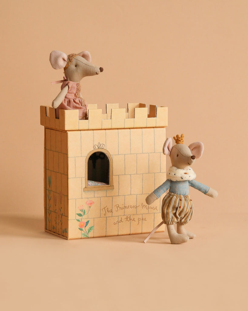 Two toy mice, dressed as prince and princess, are posed with the Maileg Princess And The Pea With Prince Set. One stands inside the floral cardboard castle box reading "The Princess Mouse and the pea" against a beige background.