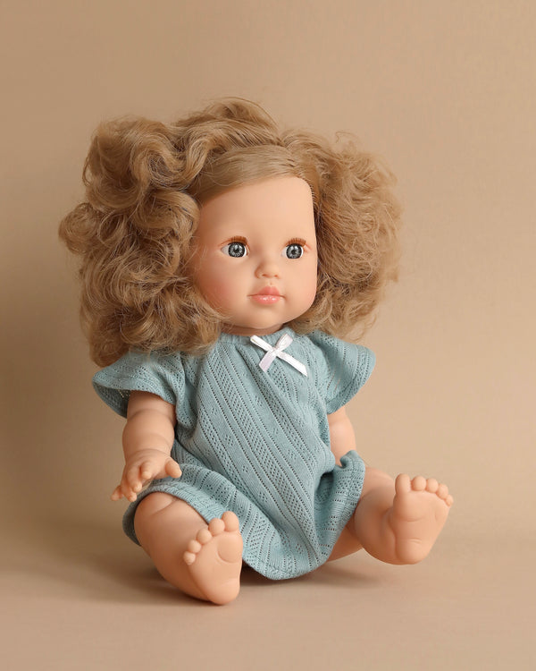 A Minikane Baby Doll (13") - Lola with curly blonde hair, wearing a light blue dress with a white bow, sits against a beige background. The doll has a calm expression with blue eyes and open, slightly outstretched arms and legs. It is anatomically correct and features a gentle, natural vanilla scent.