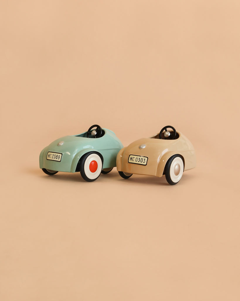 Two small toy cars from the Maileg LouiMax and Stella Adventure Set—one light blue and one beige—are positioned side by side on a beige background. Both feature white wheels with black and red details, along with visible front license plates, making them perfect for your Maileg mice to take Little Brother and Sister Mice on delightful adventures.