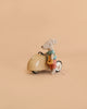 A Maileg Little Brother Adventure Set features a Little Brother Mouse dressed in a blue shirt and red shorts, sitting on a cream-colored Maileg Car and holding a yellow piece of cheese, set against a plain beige background.