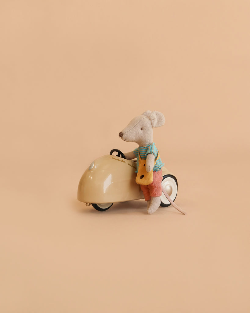 A Maileg Little Brother Adventure Set features a Little Brother Mouse dressed in a blue shirt and red shorts, sitting on a cream-colored Maileg Car and holding a yellow piece of cheese, set against a plain beige background.