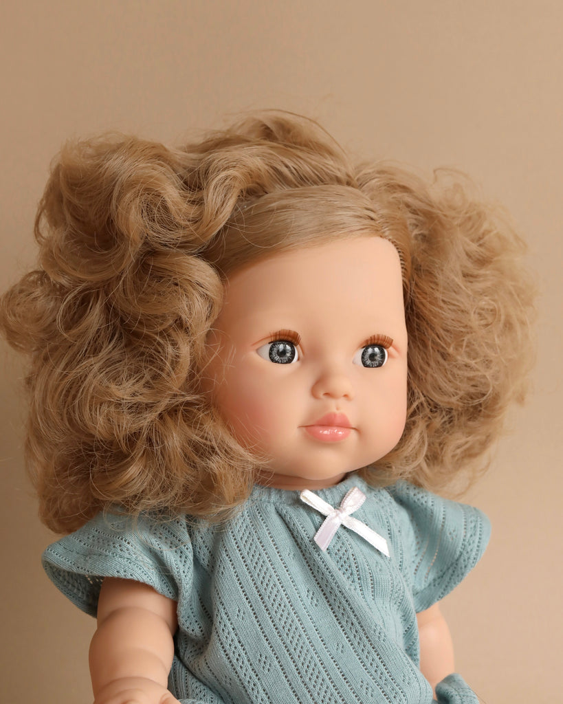 A Minikane Baby Doll (13") - Lola with curly blonde hair, blue eyes, and rosy cheeks is dressed in a light blue knitted outfit adorned with a small white ribbon. The background is a neutral beige.