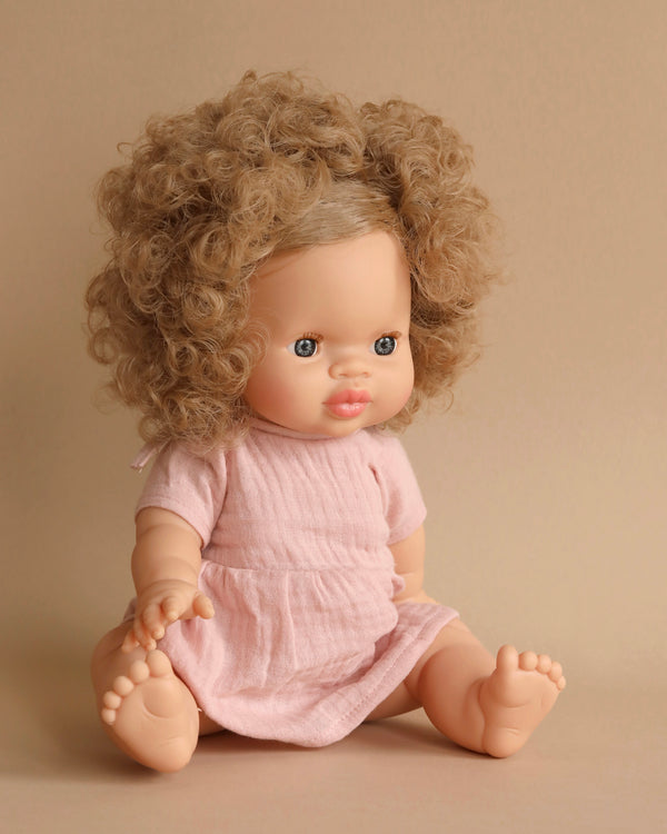 A hard-bodied doll with curly light brown hair wearing a short-sleeved, pink dress sits against a beige background. The doll has blue eyes and a calm expression. Its legs are outstretched, toes visible, resembling the lifelike Minikane Baby Doll (13") - Anais.