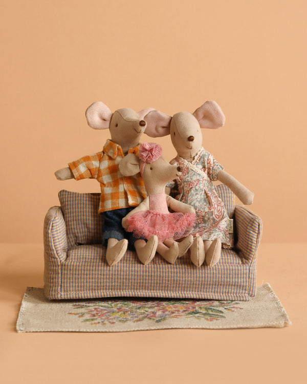 Three Maileg Family Set mice, including two adults in patterned outfits and a Ballerina Mouse in pink, sit on a small plaid sofa. They’re on a floral rug with a peach background.