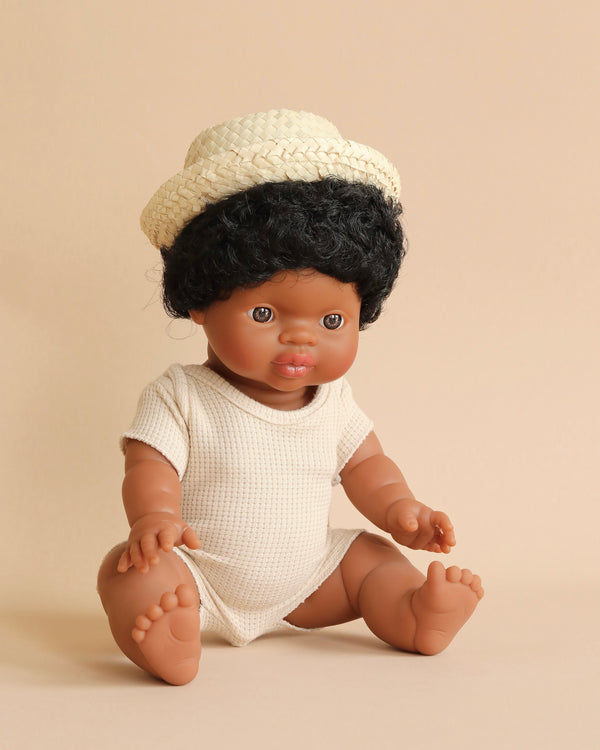 A Minikane Baby Doll (13") - Jaro with curly black hair, wearing a cream-colored onesie and a small woven hat, is seated against a neutral-colored background. The brown-skinned, anatomically correct doll has expressive brown eyes and features, with feet positioned outwards. It even has a gentle vanilla scent.