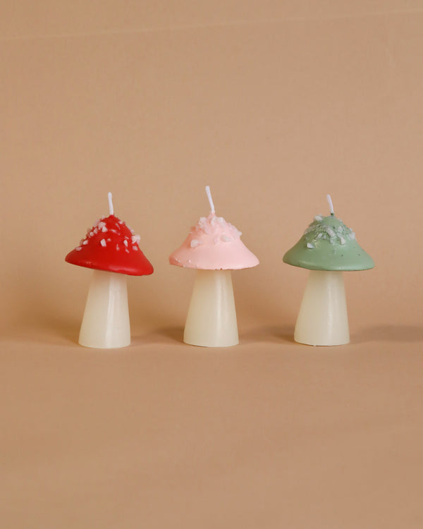 Three Meri Meri Mushroom Candles in red, pink, and green, adorned with small wax beads, displayed against a soft peach background.