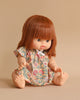 A Minikane Baby Doll (13") - Capucine with straight, auburn hair sits against a beige background. The doll is wearing a floral dress adorned with orange, green, and pink patterns, and has a white bow at the neckline. With its feet bare on a smooth surface, the doll exudes a gentle, natural vanilla scent.