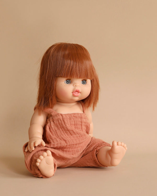 A Minikane Baby Doll With Sleeping Eyes (13") - Capucine with straight auburn hair and blue eyes, dressed in a rust-colored sleeveless jumpsuit, sits against a beige background. The anatomically correct doll's legs are splayed out in front, its hands resting on the ground, exuding a subtle natural vanilla scent.