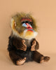 The Mandrill Monkey Puppet, a lifelike and hand-sewn toy, features vibrant colors with a striking blue and red face and a fluffy mane of multicolored fur. It sits upright against a beige background, showcasing realistic details that mimic the expression and pose of an actual mandrill.