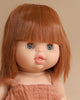 A close-up of a Minikane Baby Doll With Sleeping Eyes (13") - Capucine with straight, reddish-brown hair and bangs. The doll has round, blue eyes with freckles across its cheeks and nose, emitting a natural vanilla scent. It is dressed in a sleeveless, beige outfit. The background is a plain beige color.