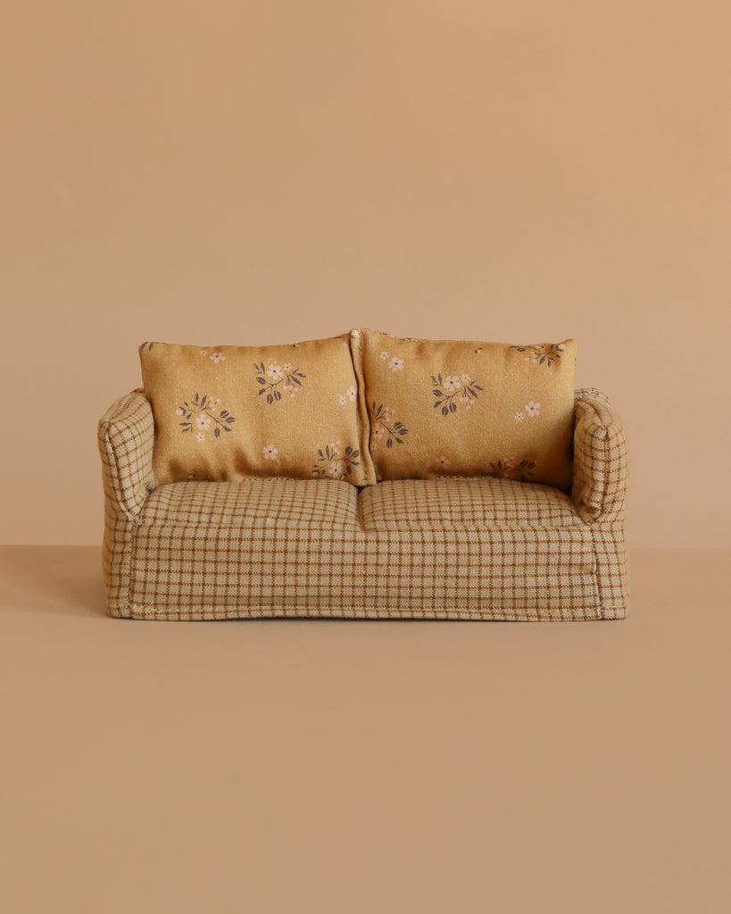 The Maileg Mom and Dad Living Room Set features a small beige plaid couch with two yellow floral cushions, placed against a light brown backdrop.