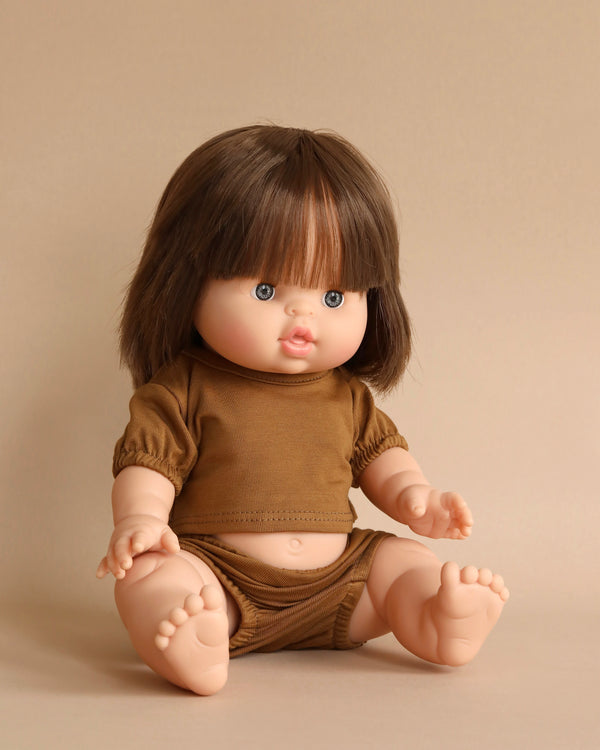 A chubby baby doll with shoulder-length brown hair sits on a beige surface. The doll, the charming Minikane Baby Doll (13") - Chloe, is dressed in a brown short-sleeved top and matching shorts. Its blue eyes and slightly open mouth give it an expressive, lifelike appearance, paired with a natural vanilla scent.
