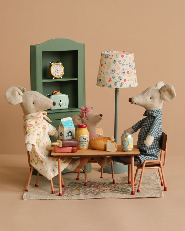 The Maileg Lunch With Grandma And Grandpa Set features three fabric mice gathered around a breakfast table, with a miniature floor lamp, green cabinet, and clock in the background for a cozy dining scene.