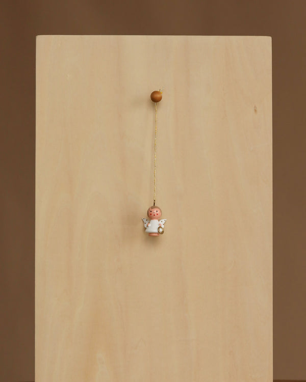 The Christian Ulbricht Ornament - Angel (White), featuring wings and a halo, is handmade in Germany and hangs by a string from the center of a plain wooden board against a neutral brown background.