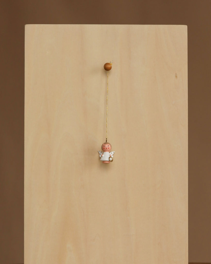 The Christian Ulbricht Ornament - Angel (White), featuring wings and a halo, is handmade in Germany and hangs by a string from the center of a plain wooden board against a neutral brown background.