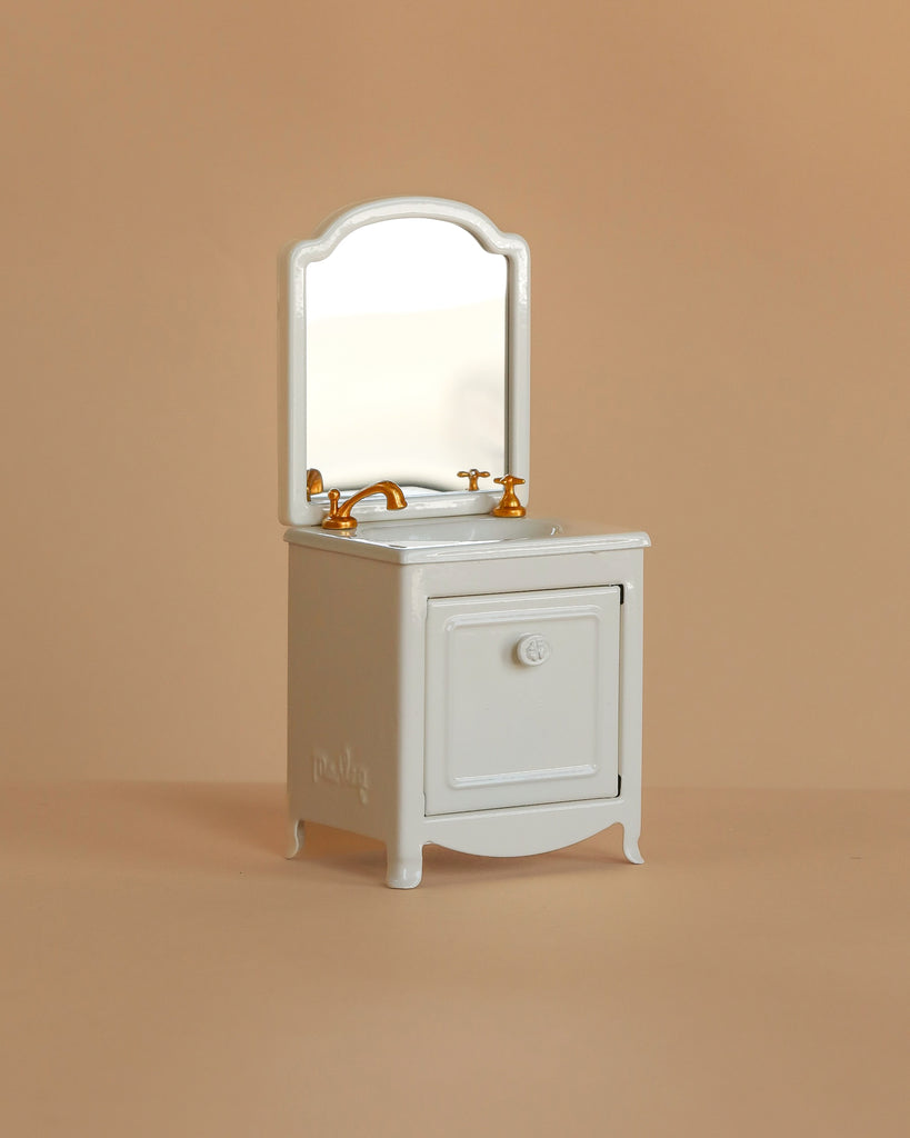 The Maileg Bathroom Set is a miniature white sink dresser with a mirrored back, gold faucets, and a single cabinet door—ideal as Maileg mouse furniture—shown against a beige background.