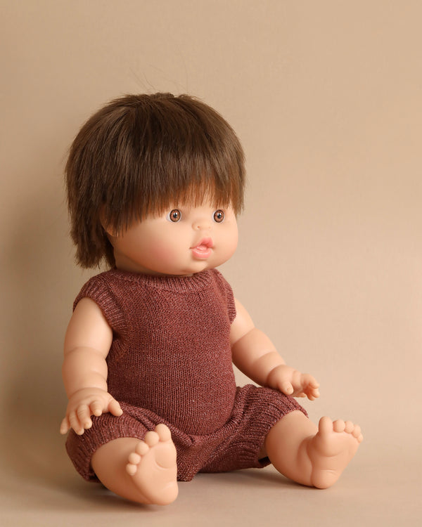 A Minikane Baby Doll (13") - Jules with short brown hair sits up against a neutral backdrop. The doll is dressed in a sleeveless, brown knit romper and has outstretched arms and bare feet. With its natural vanilla scent, the outfit and setting give the doll a simple, classic appearance.