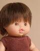 A Minikane Baby Doll (13") - Jules with short, brown hair and wide-open brown eyes is dressed in a sleeveless maroon knit top. The doll, with a light complexion and made of plastic or a similar material, has an anatomically correct design. The background is a plain, neutral color.