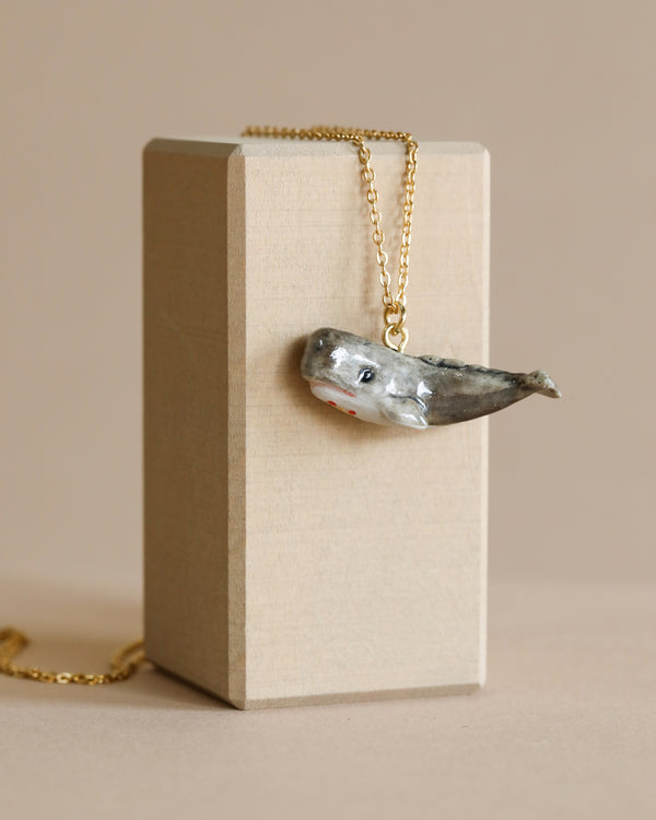 A small Whale Necklace on a 24k gold plated chain displayed on a beige rectangular jewelry box against a neutral background. The whale charm, hand painted with detailed features, has a slight hint of