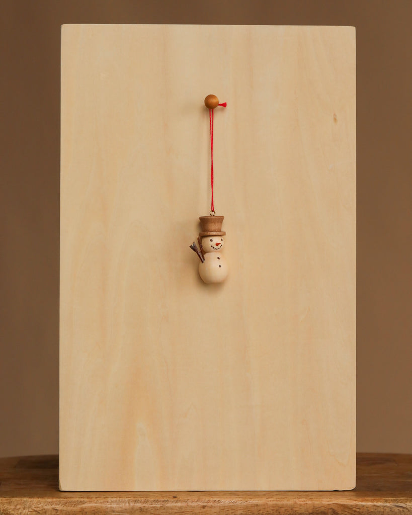The Christian Ulbricht Ornament - Snowman, made in Germany, hangs by a red string from a peg on a simple light wood board, set against a plain brown background.