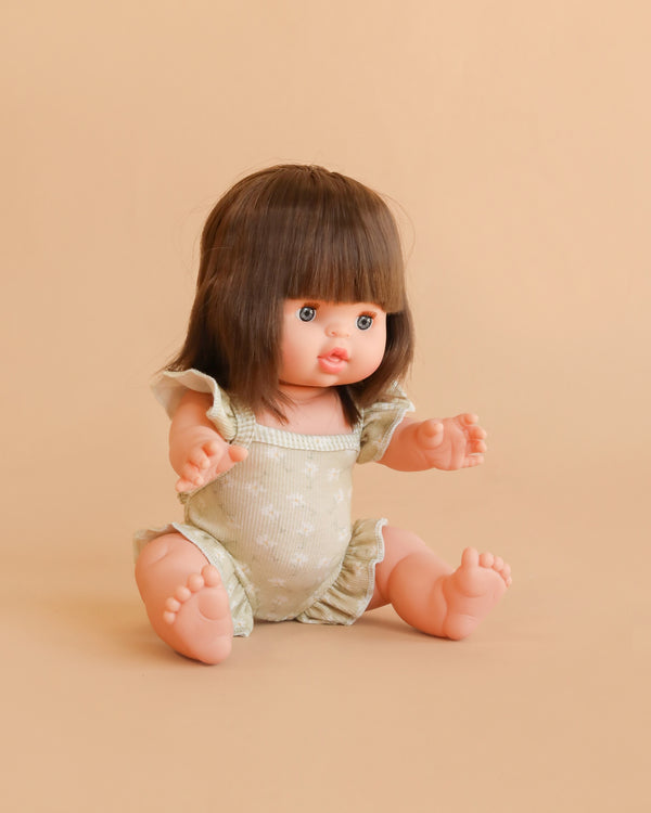 The Minikane Baby Doll (13″) - Chloe with Clothing features short brown hair and bangs, and is dressed in light-colored clothes while sitting on a beige background with arms reaching forward and legs spread apart.