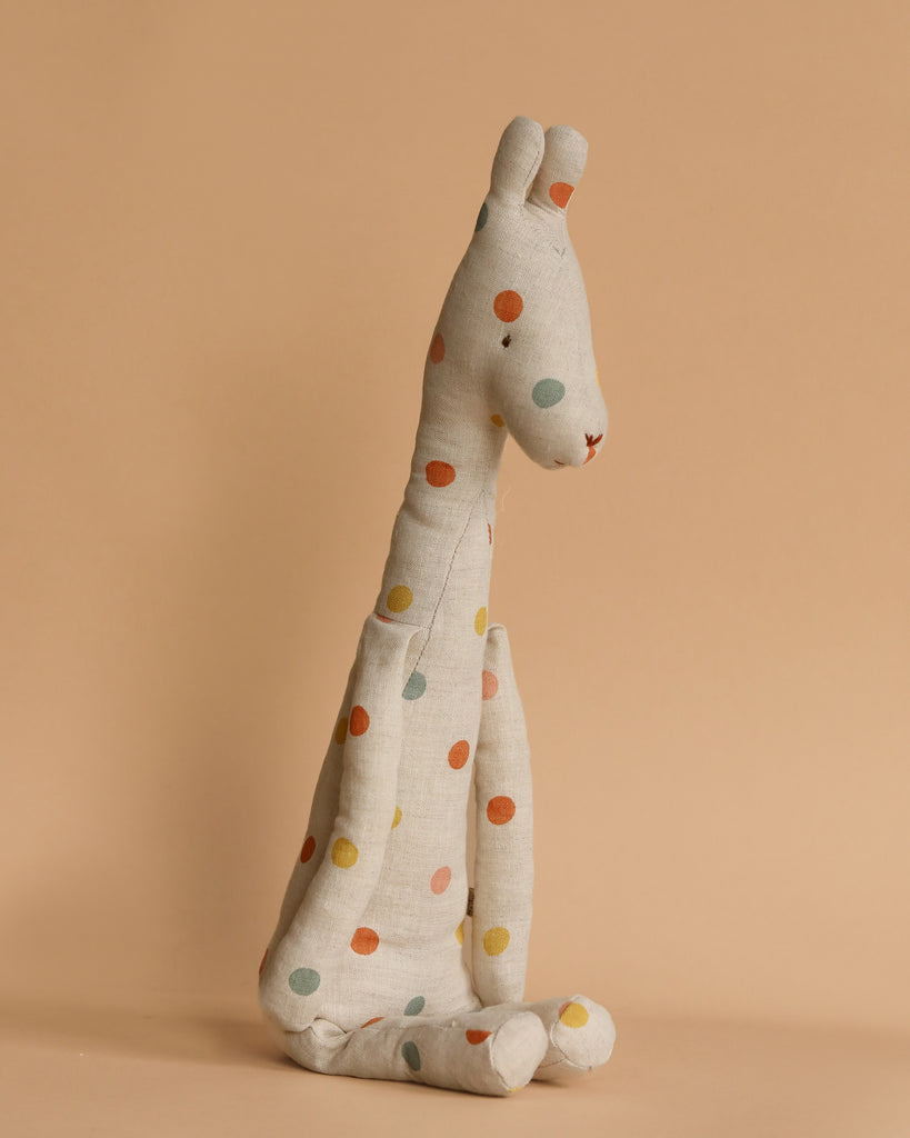 The Maileg Medium Giraffe is an upright plush toy made from recycled polyester, featuring a light beige fabric with vibrant polka dots in red, blue, yellow, and green. This safari companion rests beautifully against a warm beige backdrop.