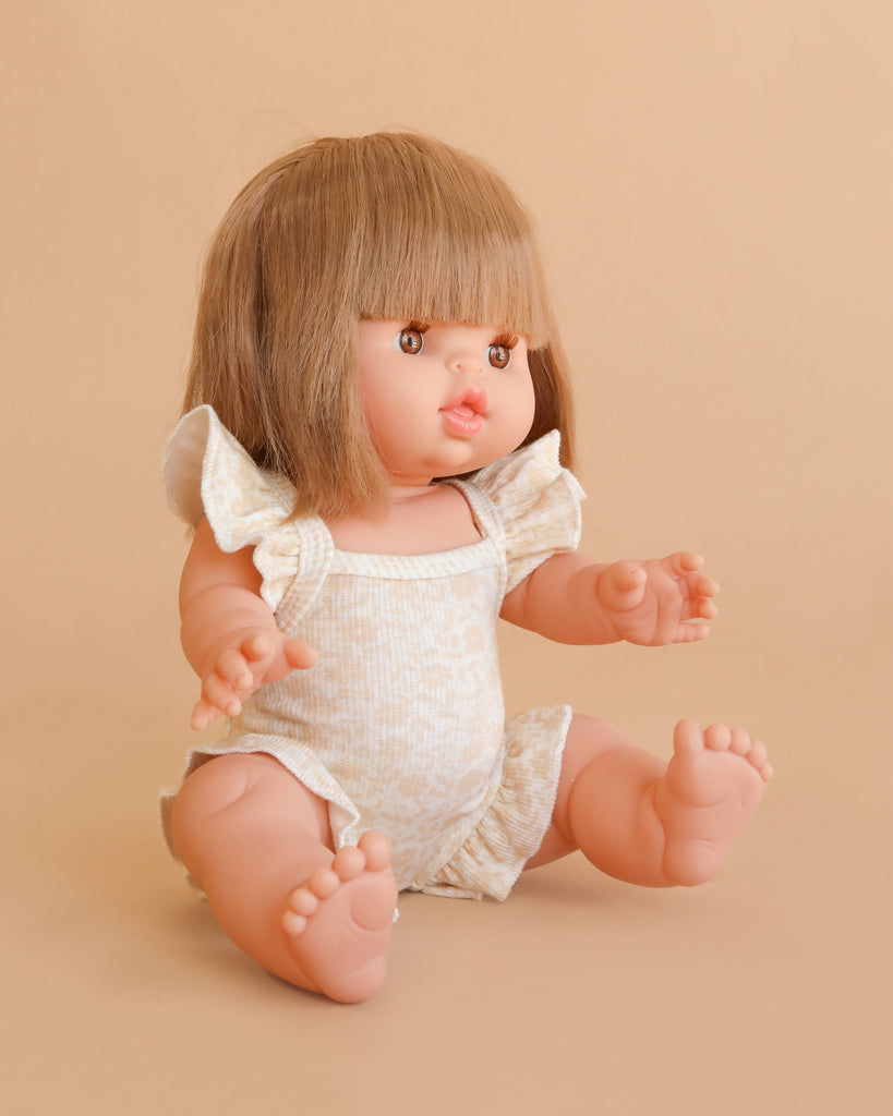 A Minikane Baby Doll (13″) - Zoe with Clothing, featuring brown hair with bangs and a light patterned romper, sits on a beige background with arms and legs outstretched.