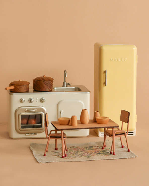 The Maileg Natural Kitchen Set includes a vintage fridge, cream stove and sink, two brown pots, dining table with dishes, two chairs, and a small rug—perfect for any handmade wooden dollhouse.
