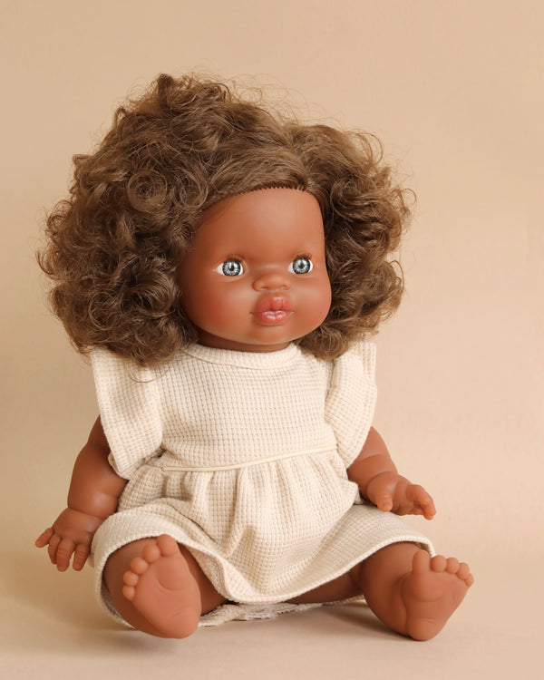 A seated Minikane Baby Doll (13") - Charlie with curly brown hair and blue eyes, wearing a white, short-sleeved dress. The doll has a light brown complexion and is set against a plain beige background, emanating a subtle vanilla scent.