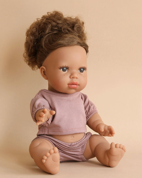 A Minikane Baby Doll (13") - Melissa with curly brown hair sits against a beige background. The doll, dressed in a short-sleeved, light purple top and matching bottoms, has a lifelike appearance with detailed facial features and visible toes and fingers. It even has an anatomically correct design for added realism.