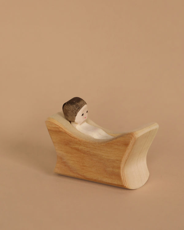 A small wooden figurine, the Ostheimer Baby Jesus with brown hair, is lying in a curved wooden bathtub against a plain beige background, embodying the charming simplicity characteristic of handcrafted wooden toys.