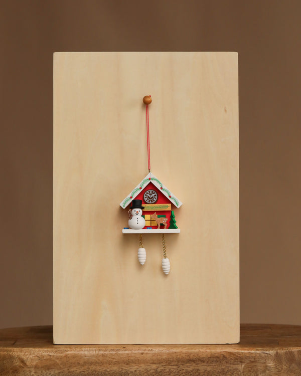 The Christian Ulbricht Ornament - Snowman with Red Cuckoo Clock, made in Germany, features a snowman and pine tree and hangs against a plain light wooden background on a centered wooden surface.